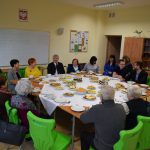 Grupa seniorów siedzących przy stole wraz z Wójtem Gminy Puławy, pracownikami GOPS-u i biblioteki.