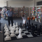 Seniorzy grają w szachy na rozłożonej na podłodze wielkiej szachownicy.
